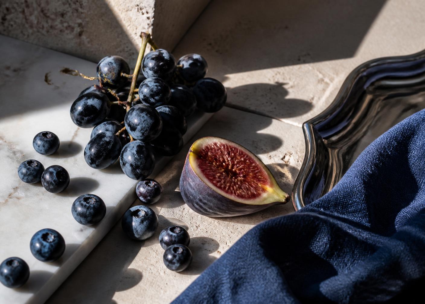 Blue grapes, blueberries and a halved fig on travertine and marble in hard sunlight